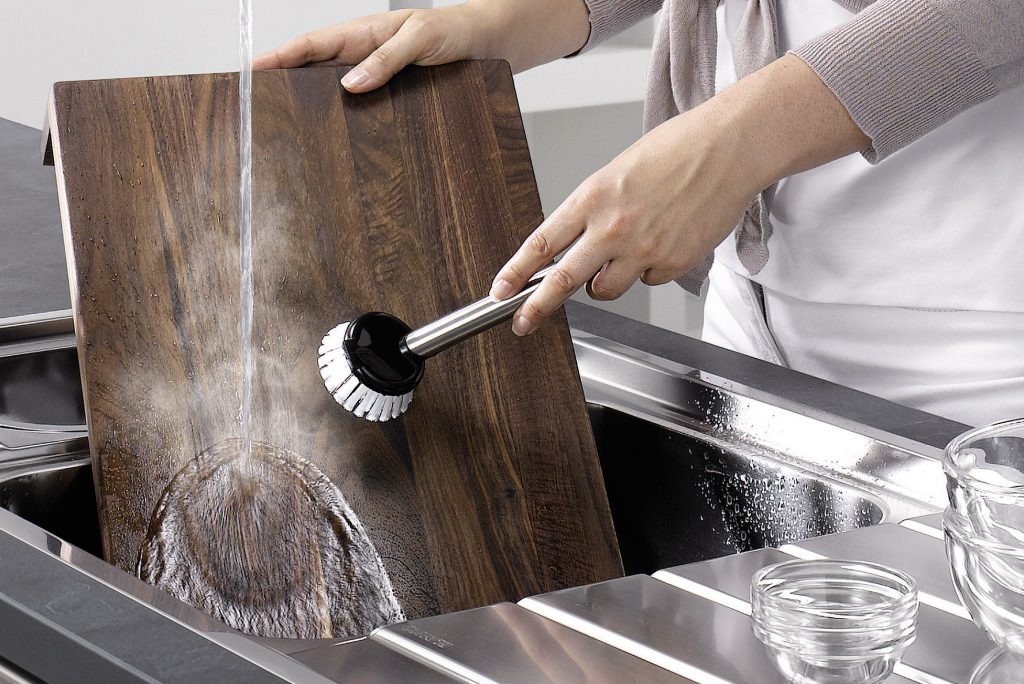 A dark cutting board is cleaned in a stainless steel sink